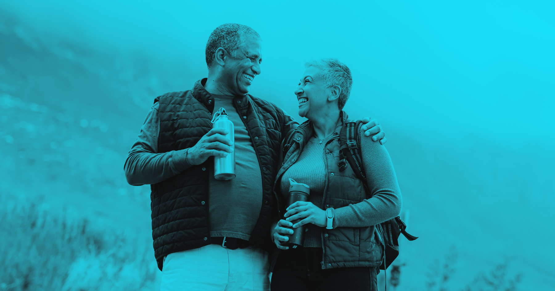 A man and woman are walking in the hilly countryside. The man has his arm around the woman and they are smiling at one another.