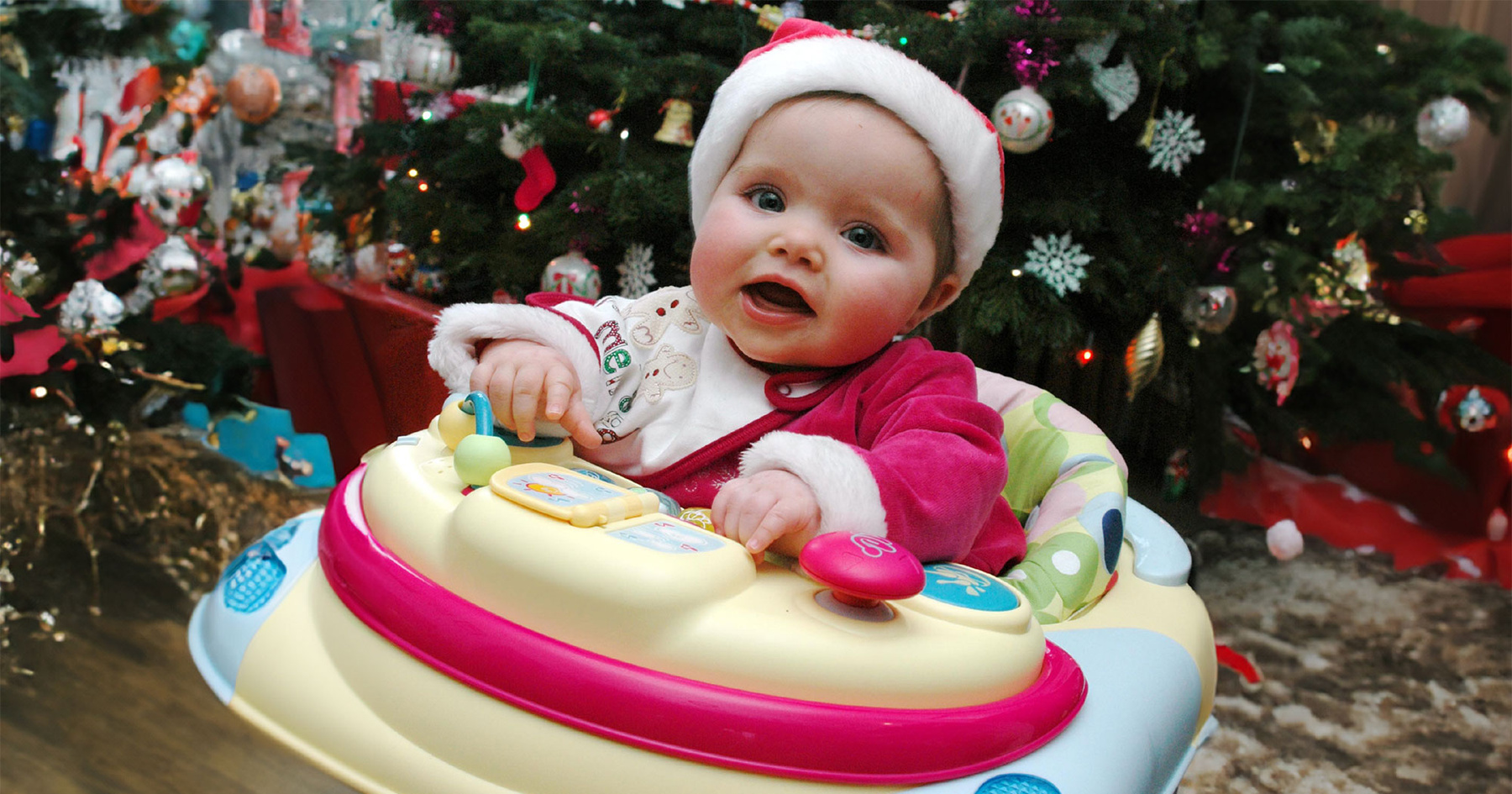 A happy baby in a Santa hat sits in a colourful walker in front of a decorated Christmas tree