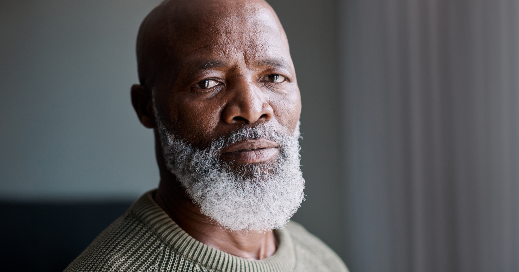 Elderly man with a grey beard and serious expression, wearing a green sweater, against a blurred background