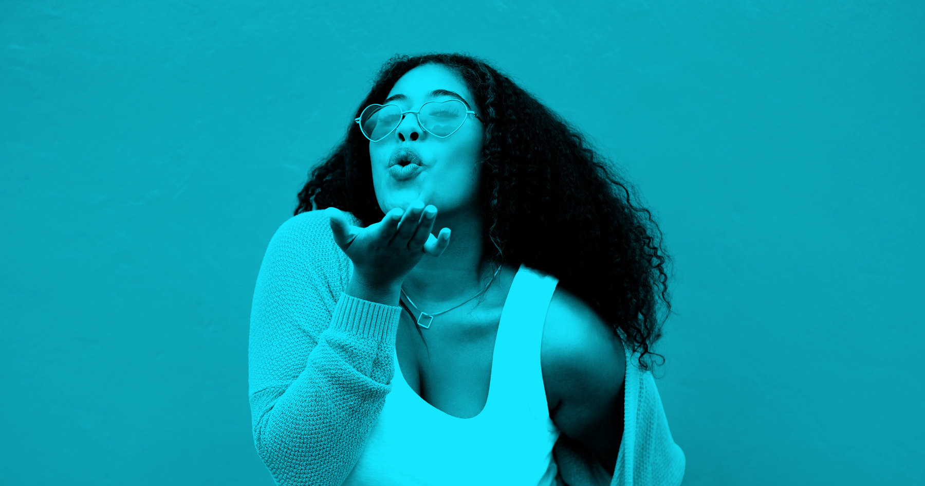 A young woman with curly hair wearing heart-shaped sunglasses, a white tank top, and a cardigan blows a kiss toward the camera