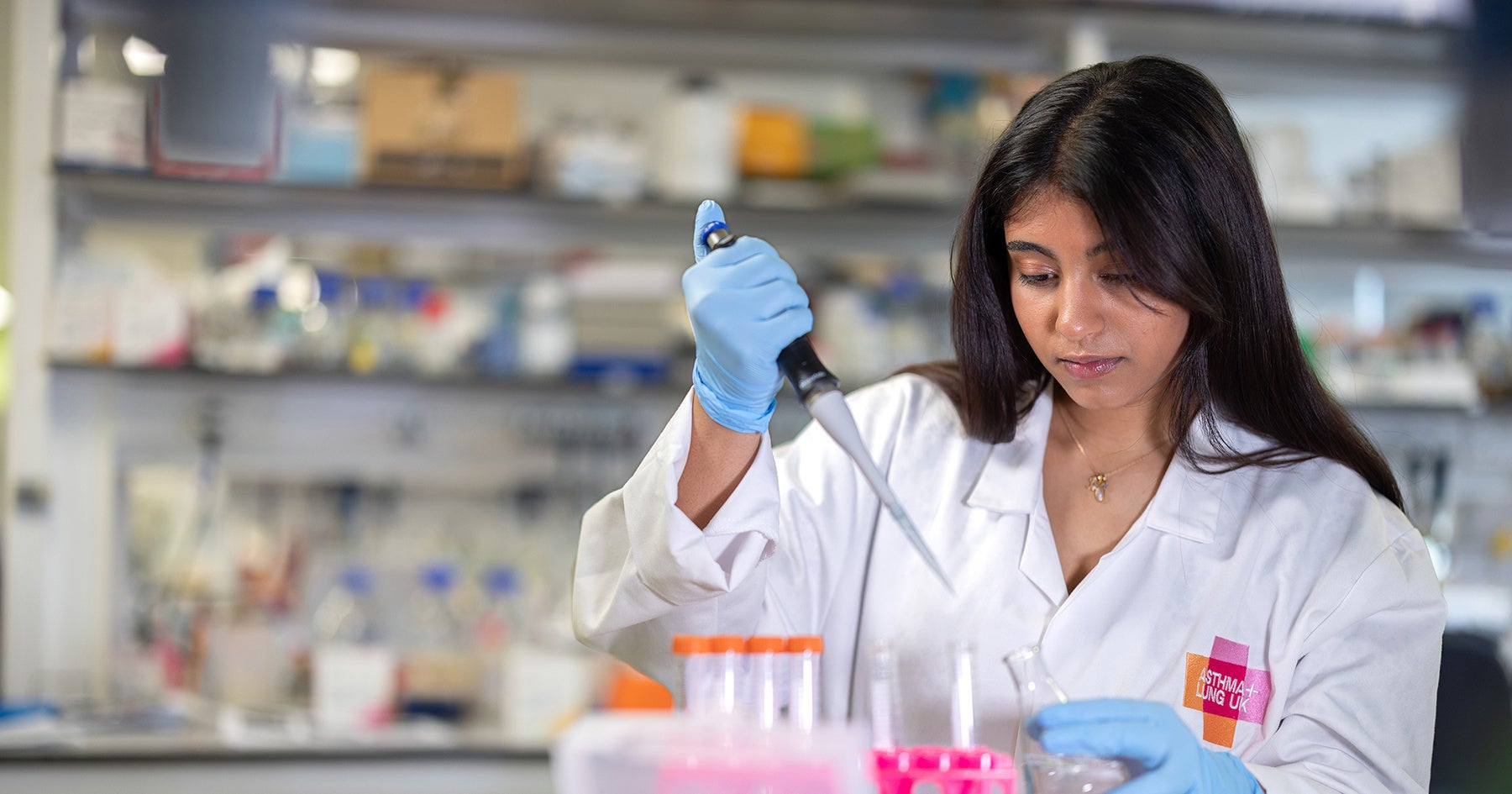 Researcher in a lab coat uses a pipette to transfer liquid into a flask at a laboratory bench