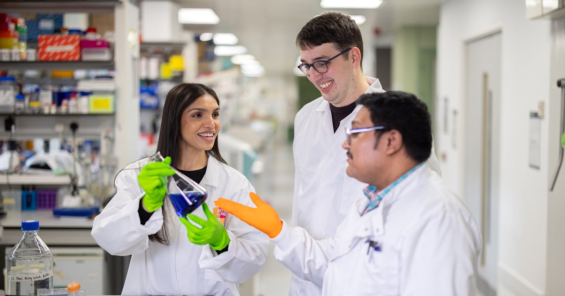 Three researchers in lab coats talk and smile in a laboratory, one holding a flask with blue liquid