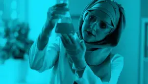 A lady is in a lab, wearing a lab coat, plastic gloves and goggles. She is holding up a beaker of liquid and looking straight at it.