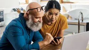 A couple are using a laptop. One is also referring to their phone. They are in the kitchen, and the laptop is on the countertop.