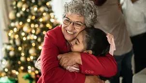 User An elderly woman with grey curly hair, wearing glasses and a red cardigan, hugs a young girl in front of a lit Christmas tree
