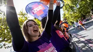 A woman in a purple shirt with raised arms cheering with pom-poms at a sporting event