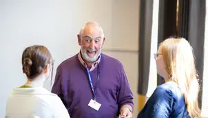 Three people in conversation, a man in a purple sweater talking to two women