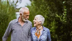 An elderly couple smiling at each other, walking outdoors with greenery in the background