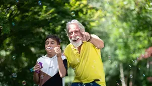 A boy and a senior man playfully blowing bubbles in a sunlit  garden