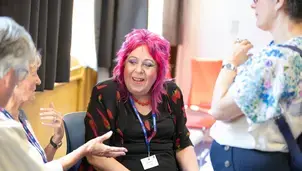 A person with vibrant pink hair engaging in conversation with two others, gestures suggesting an animated discussion