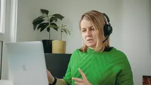 A woman with a headset looks at her laptop, with a plant in the background
