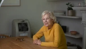  An elderly woman with a nasal oxygen tube sits calmly at a table, with glasses in front of her and shelves in the background