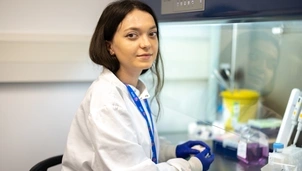 A woman with long dark hair wearing a white lab coat is sat in a lab looking directly to camera