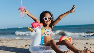 A young girl wearing white framed sunglasses and a colourful bikin is on a beach sat in a unicorn inflatable with her arms out smiling.