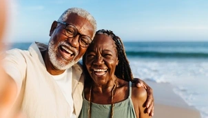 A man with short white hair and a beard and a woman with long braided hair are stood on a beach hugging and smiling at the camera while taking a selfie.