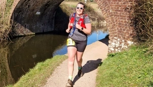 A woman with dark hair tied back is running alongside a canal on a sunny day. She's wearing a grey t shirt, black shorts and a red running vest.
