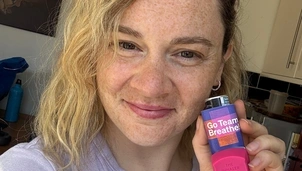 A woman with short blonde hair is wearing a lilac t shirt and holding up an Asthma + Lung UK branded inhaler smiling at the camera