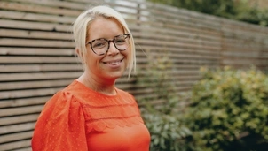 A woman with short blonde hair and glasses is wearing an orange top. She's stood outside in a garden looking directly to camera