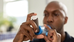 A man wearing a black polo shirt is holding a brown inhaler with a spacer. His hands and inhaler are close up and his face and the background are out of focus.