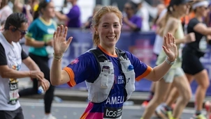 A woman with ginger hair in a pony tail is wearing an Asthma + Lung UK t shirt running the London Marathon. She's looking directly at the camera smiling and waving and there are other runners in the background.