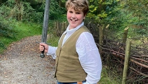 A young boy with light brown hair is stood on a country path wearing blue jeans, a light blue shirt, brown waistcoat and black and white trainers. He is smiling at the camera and holding up a toy sword.