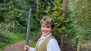 A young boy with light brown hair is stood on a country path wearing blue jeans, a light blue shirt, brown waistcoat and black and white trainers. He is smiling at the camera and holding up a toy sword.