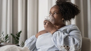 A woman with mid length afro hair is sat down on a chair in front of cream curtains. She's wearing a blue and white striped blouse and she is using an oxygen mask and has her hand on her chest.