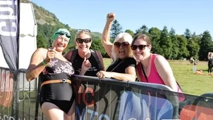 A group of women stood in a fiedl on a sunny say. one of them is wearing a black swimming costume and a blue swimming hat smiling and giving the thumbs up.