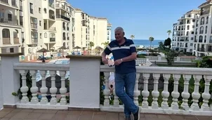 A man with short grey hair, a blue and white polo shirt and blue jeans is stood in front of a railing with the sea in the background.