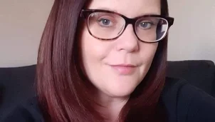 A woman with shoulder length dark hair wearing glasses and a dark top is sat on a sofa in a living room and looking directly at camera.