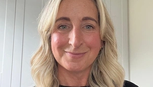 A woman with mid length blonde hair is stood inside wearing a black t shier and smiling at the camera