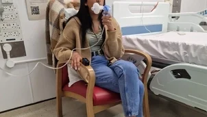 A woman with long dark hair is wearing blue jeans, black converse and a beige cardigan is sat in a hospital chair next to a hospital bed with an oxygen mask.
