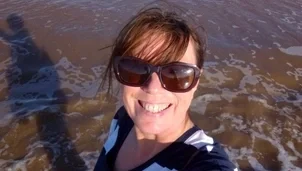 A woman with long dark hair in a pony tail, dark sunglasses and a black and white striped top is stood in the sea taking a selfie and smiling to the camera