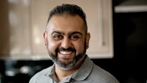 A man with short dark hair and a dark beard is wearing a grey polo shirt and smiling to camera