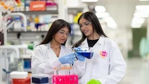 Two female scientists in lab coats and safety glasses work together in a laboratory, pouring blue liquid from a flask into test tubes