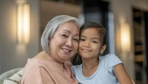 An elderly woman and a young girl smile warmly, sitting close together indoors