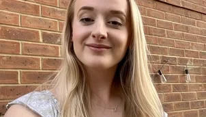 A woman with long blonde hair wearing a grey top standing in front of a brick wall and smiling to camera.