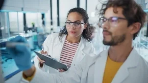 Two scientists in lab coats and safety glasses work together in a laboratory, one writing on glass while the other takes notes