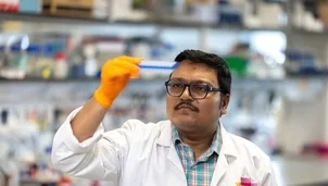 Scientist in lab coat and orange glove examines a test tube rack in a laboratory