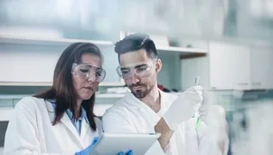 Two scientists in lab coats and safety glasses review data on a tablet in a laboratory