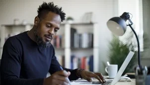 A man writes notes while working on a laptop at a desk