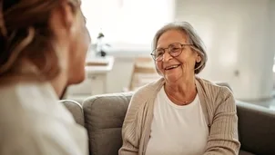 An elderly woman with glasses smiles while talking to another person indoors