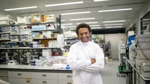 A smiling scientist in a white lab coat stands with arms crossed in a laboratory