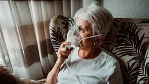 An older woman sits in a chair using an oxygen mask