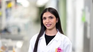 A researcher in a white lab coat with an Asthma + Lung UK logo stands smiling in a laboratory