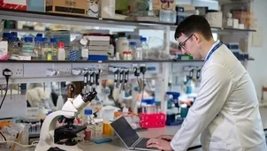 A scientist in a lab coat works on a laptop at a laboratory bench with equipment and supplies