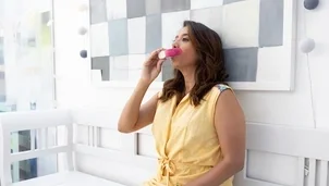 A woman in a yellow dress sits on a white bench using a pink inhaler