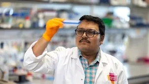 A man with short dark hair and a dark moustache is wearing black glasses, a white lab coat and orange gloves. He's stood in a research lab holding some samples up to the light