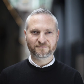 Man with short grey hair and beard wearing a black jumper over a white shirt, looking at the camera