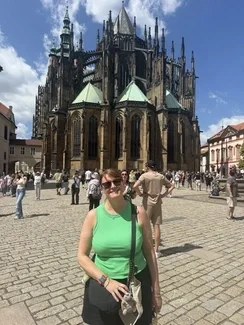 A woman in a green sleeveless top and sunglasses smiles in front a cathedral, with tourists walking around the cobblestone square